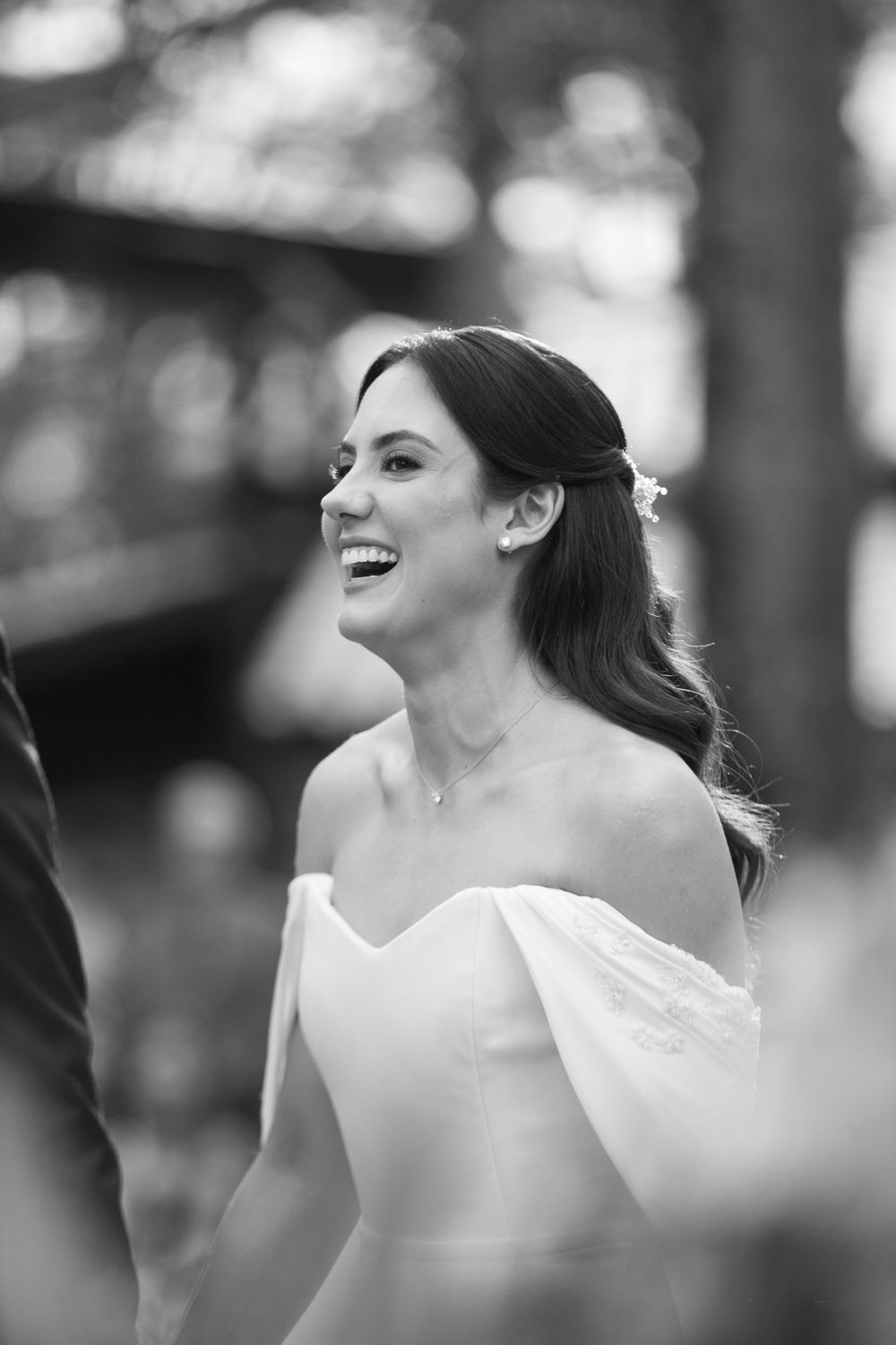 Black and white photo of a smiling bride in an off-the-shoulder gown with a floral hairpiece laughing during an outdoor wedding ceremony.