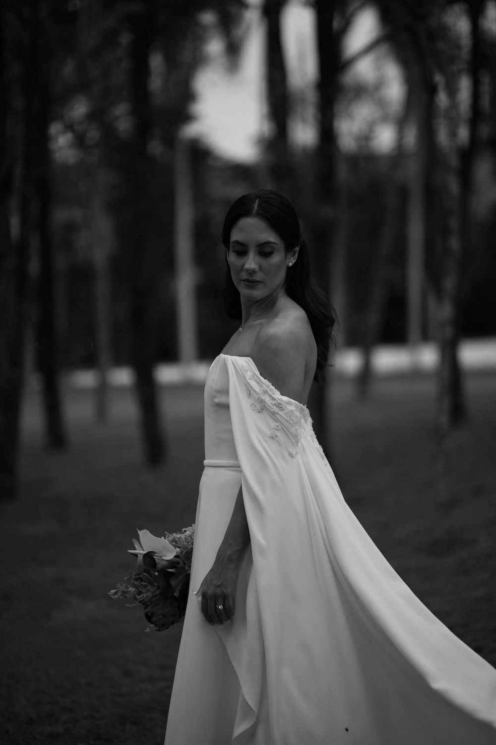 Black and white portrait of a bride in a flowing off-the-shoulder gown holding a bouquet among tall trees.