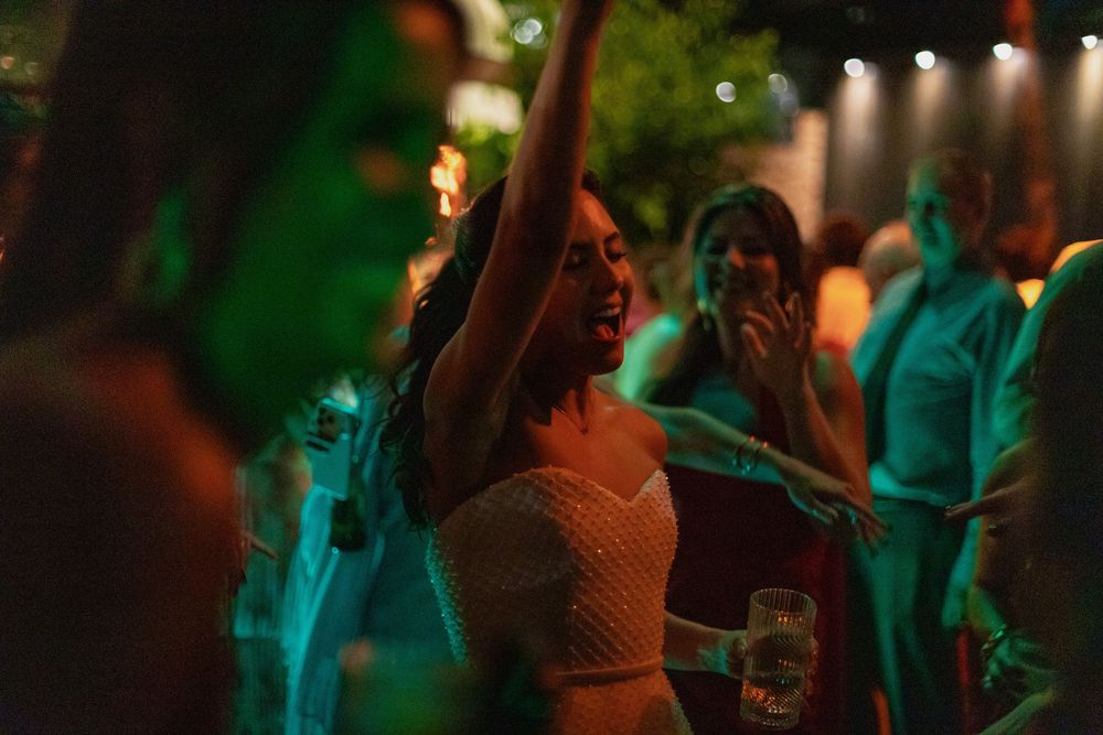 Woman in a lace dress dances with one arm raised at a lively evening party while guests laugh and celebrate under warm string lights.