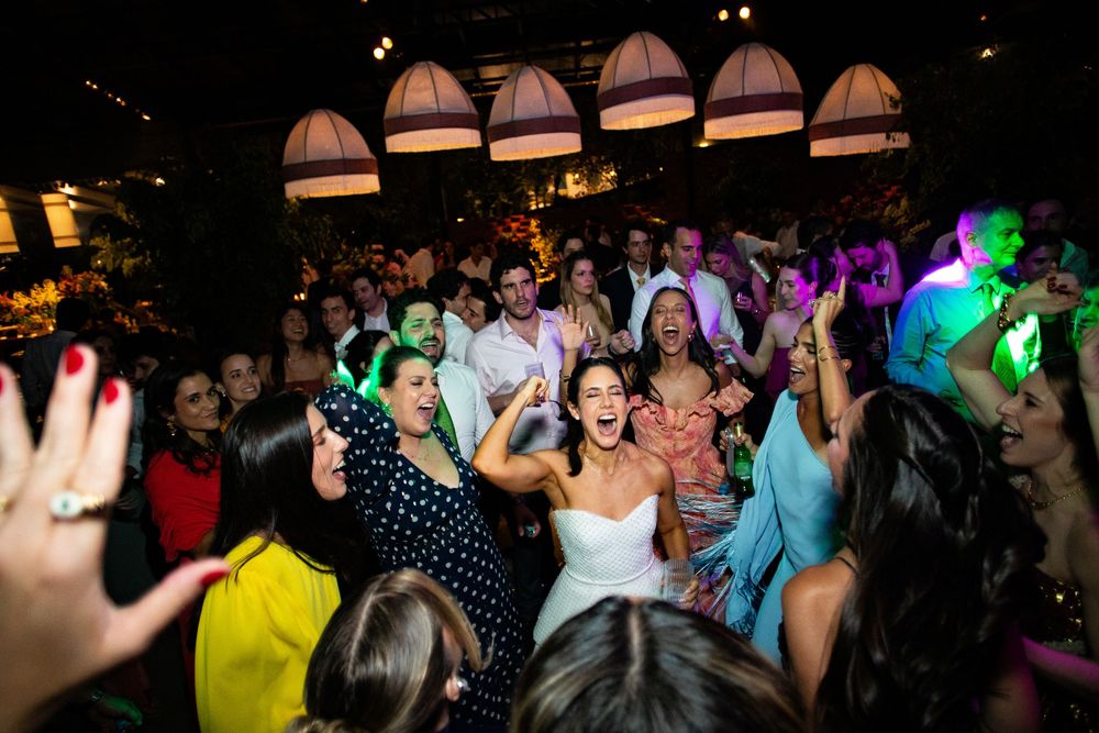 Joyful wedding guests dance and cheer on a crowded dance floor beneath warm pendant lights, with a bride in a white dress at the center.