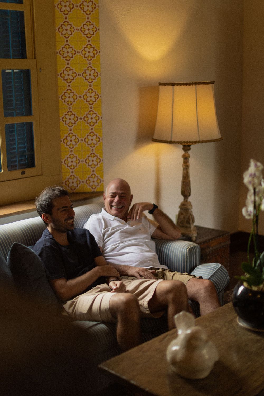Two smiling men in casual clothes relax together on a striped sofa in a warmly lit room with a table lamp and yellow patterned tile wall.