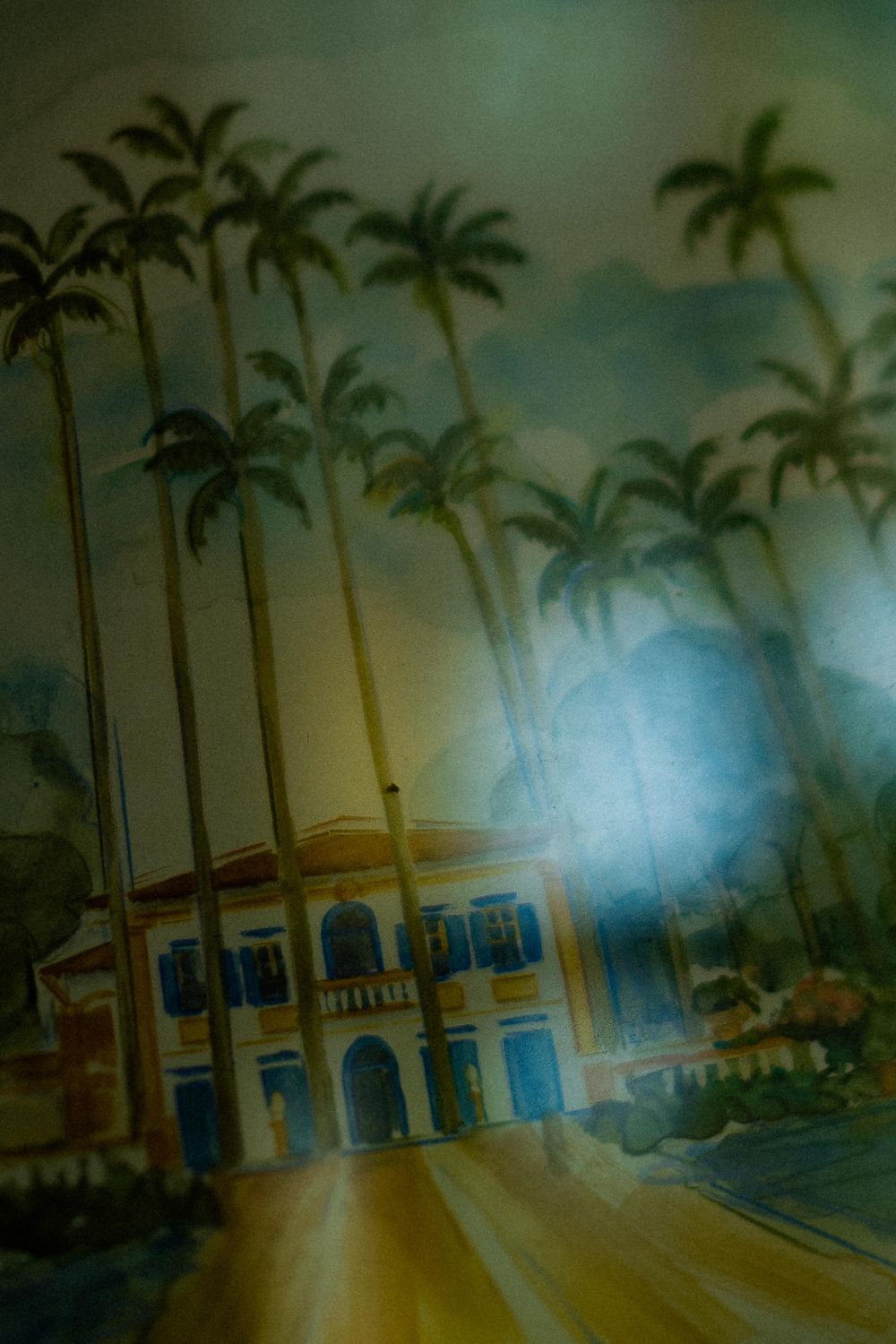 Watercolor painting of a yellow colonial building with blue shutters framed by tall royal palm trees under a hazy, misty sky.