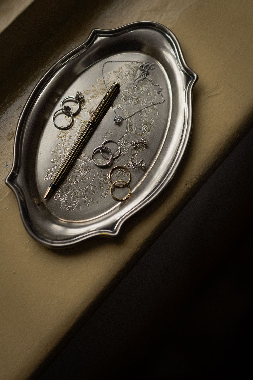 Ornate engraved silver tray holding a fountain pen, gold rings, and delicate diamond jewelry in dramatic, moody window light.