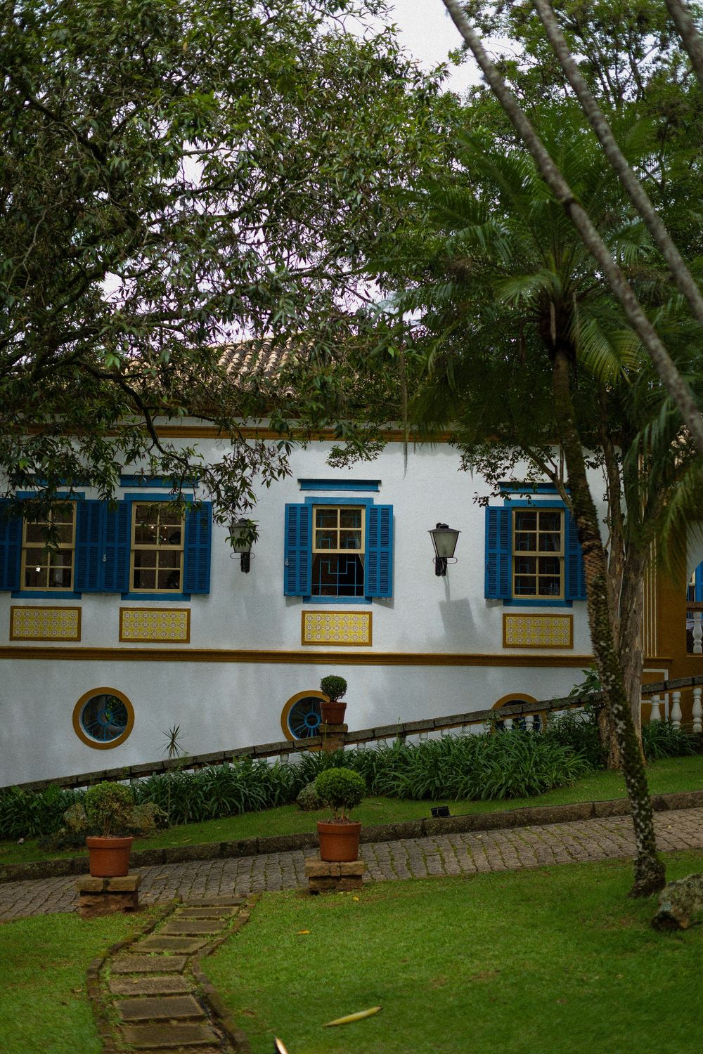 White colonial-style house with blue shuttered windows, round portholes, terracotta pots and a stone path through a lush green garden.