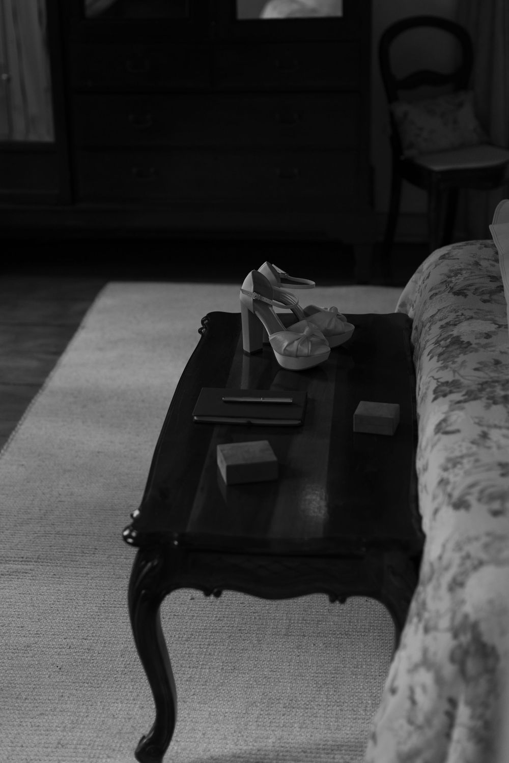 Black and white photo of platform high-heeled sandals, a clutch and small boxes on a dark vintage coffee table beside a floral bed.
