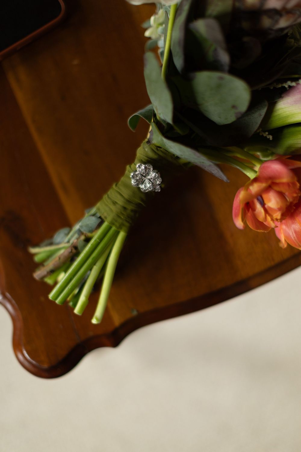 Wedding bouquet with eucalyptus leaves, a coral tulip and green stems wrapped in twine with a jeweled brooch, resting on a wooden table.