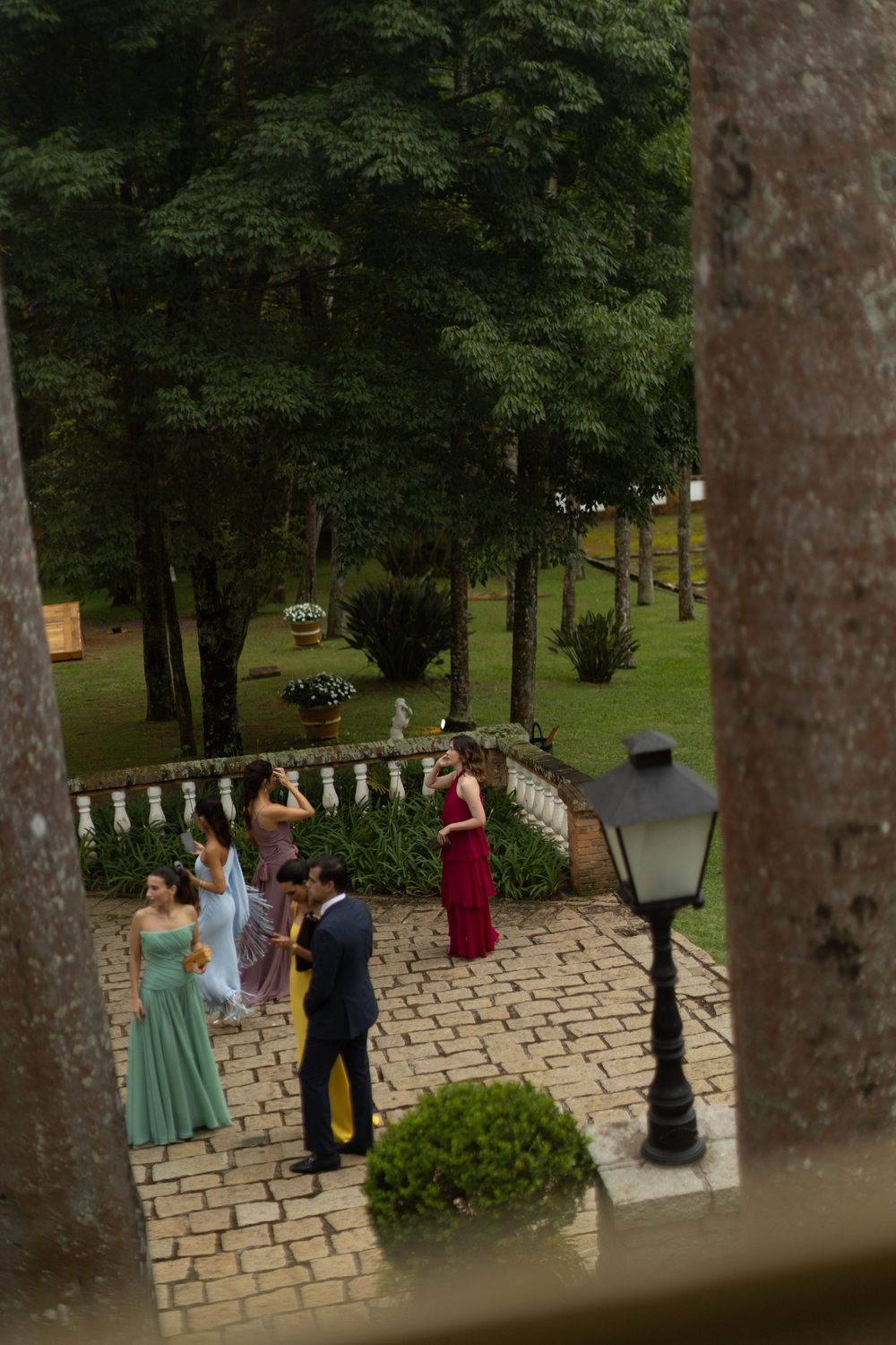 Elegant wedding guests in colorful gowns and a suit gather on a brick garden patio beside a vintage lamp post and lush green trees.