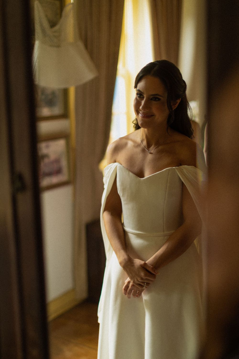 Smiling bride in an elegant off-the-shoulder white wedding gown with cape sleeves, standing in a softly lit room with hands clasped.