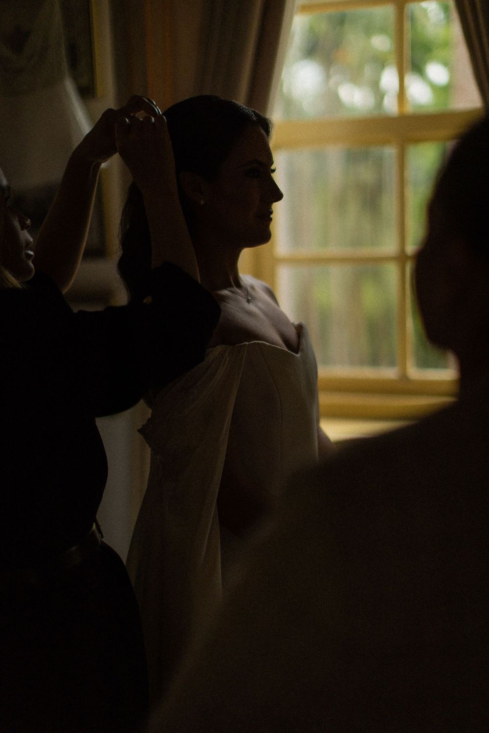 Bride in profile in a softly lit room by a window as helpers adjust her hair and gown before the wedding ceremony.