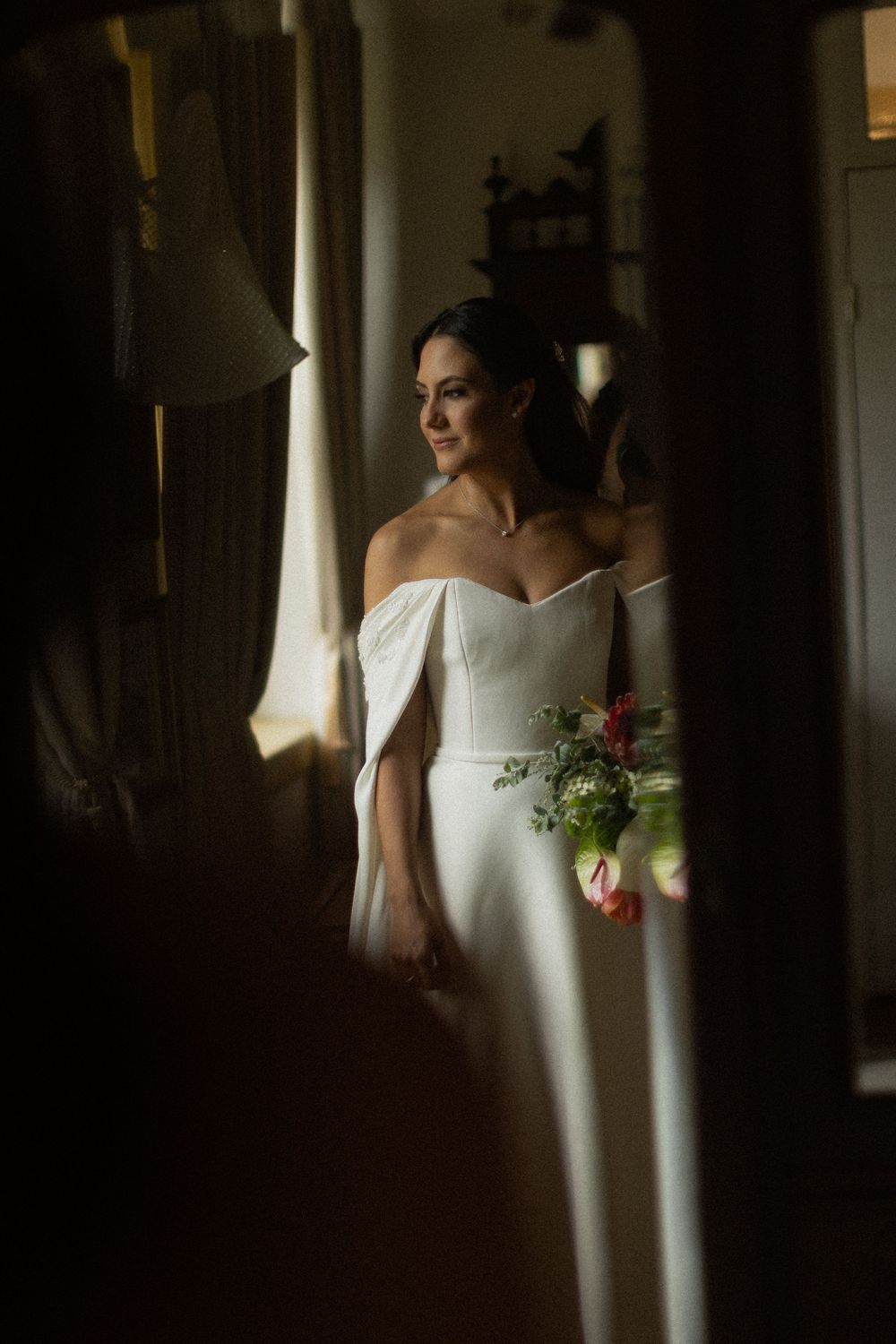 Bride in an elegant off-the-shoulder white wedding gown holds a bouquet while gazing out a window in soft, moody natural light.
