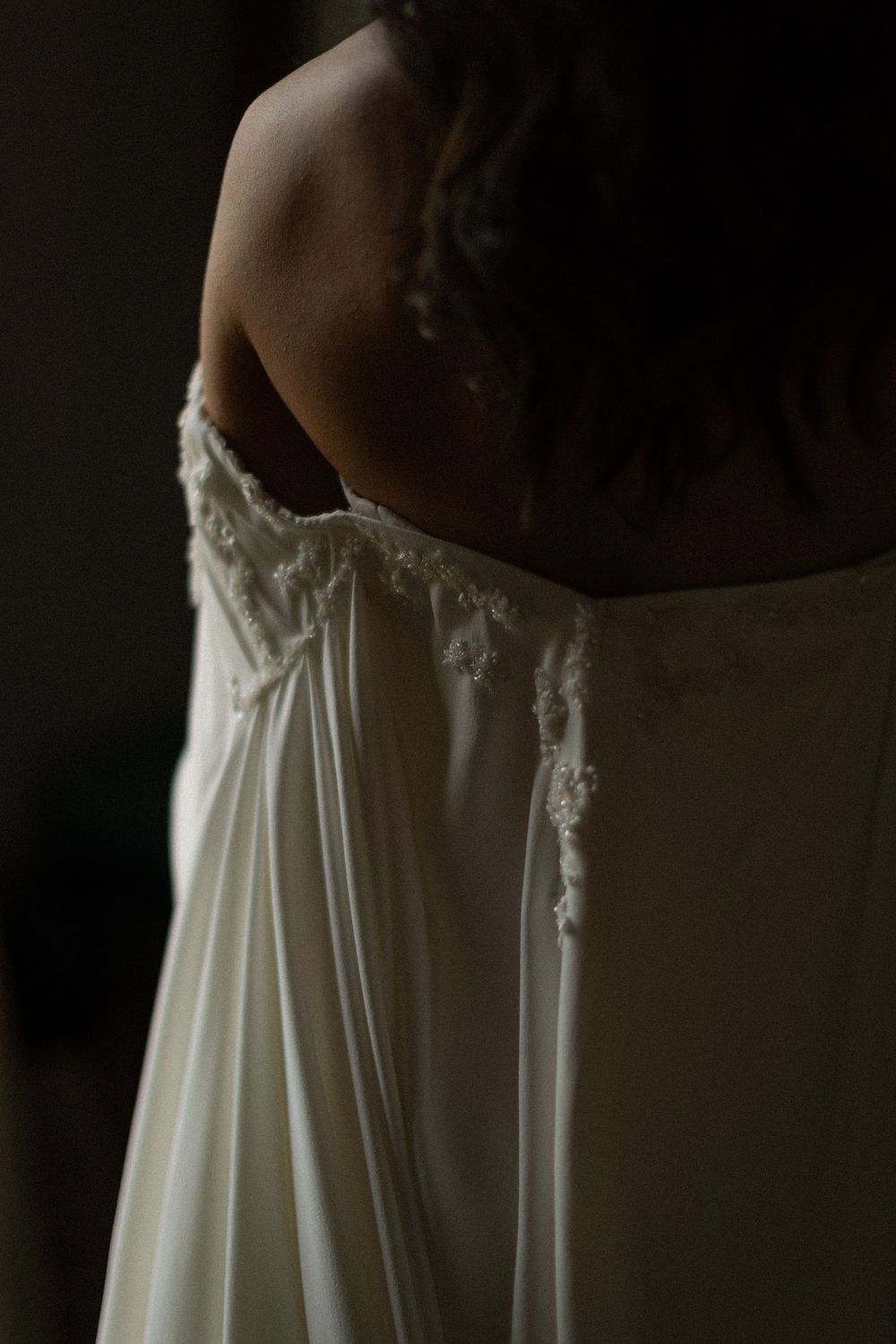 Bride seen from behind in a strapless ivory wedding gown with delicate beaded lace detailing, softly lit against a dark background.