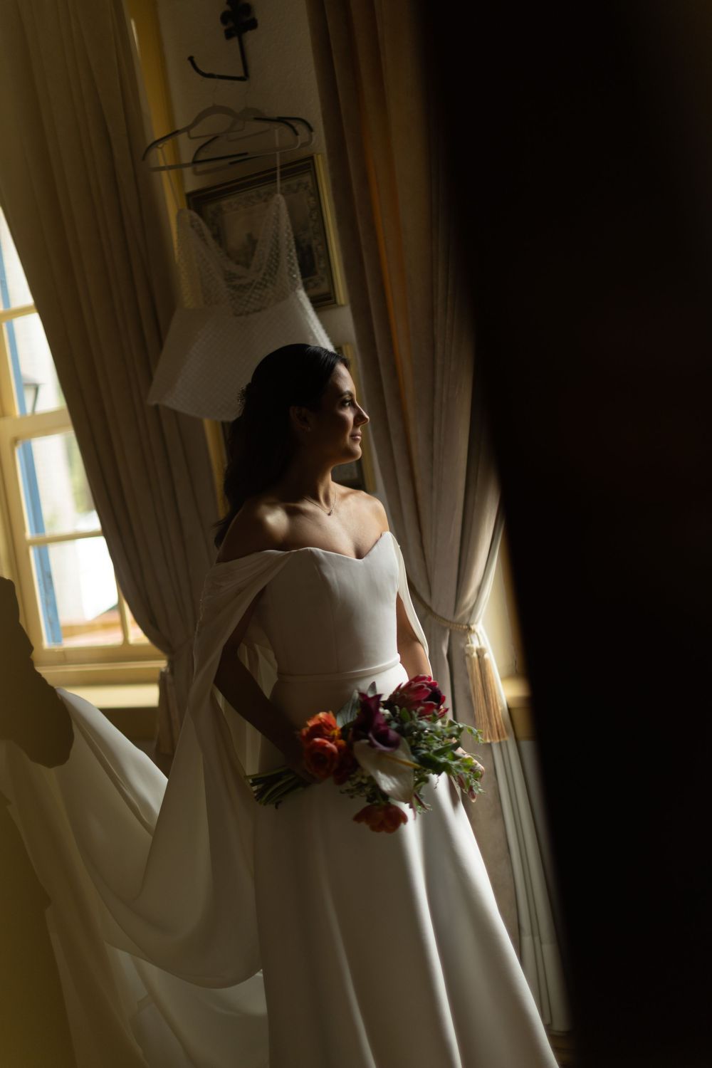 Bride in an elegant off-the-shoulder white gown holds a colorful bouquet while gazing out a sunlit window, with a dress hanging nearby.
