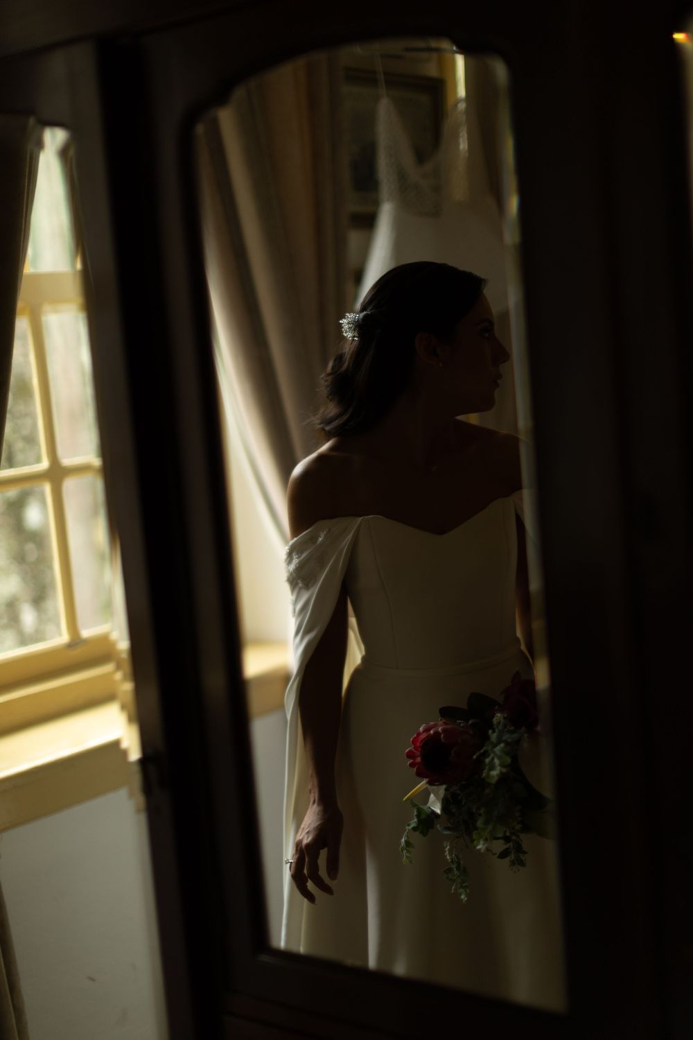 Bride in an off-the-shoulder white wedding gown holding a bouquet of red roses, reflected in a mirror beside a sunlit window.