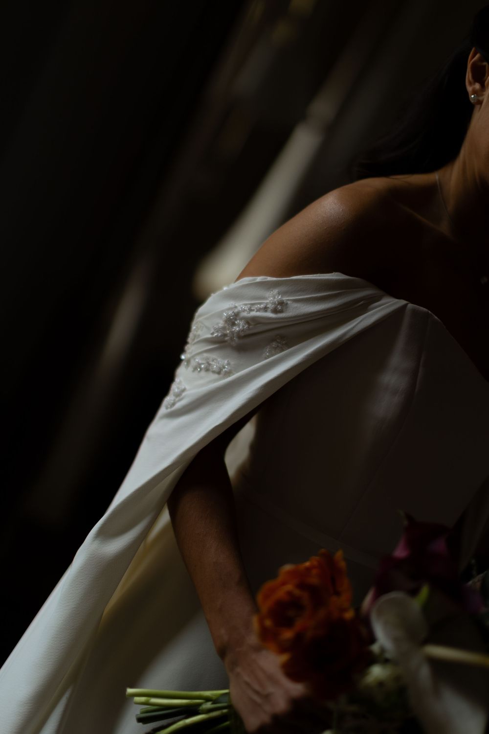 Bride in an off-the-shoulder white satin gown with beaded detail holding an orange and burgundy bouquet in dramatic, moody light.