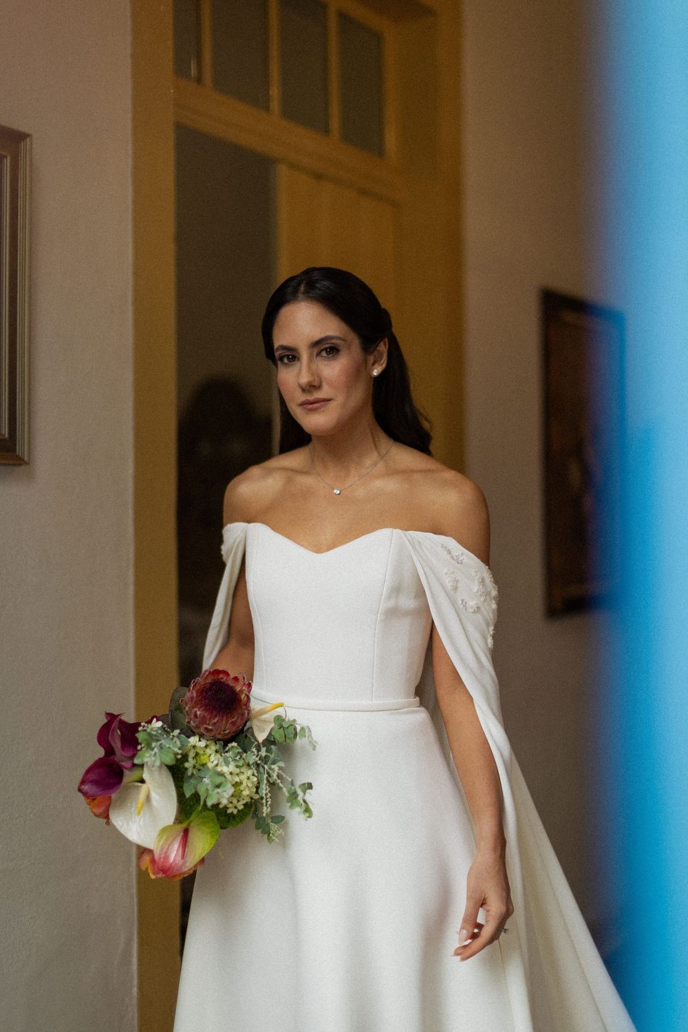Bride in an off-the-shoulder white gown with cape sleeves holds a colorful calla lily bouquet while standing in a softly lit doorway.