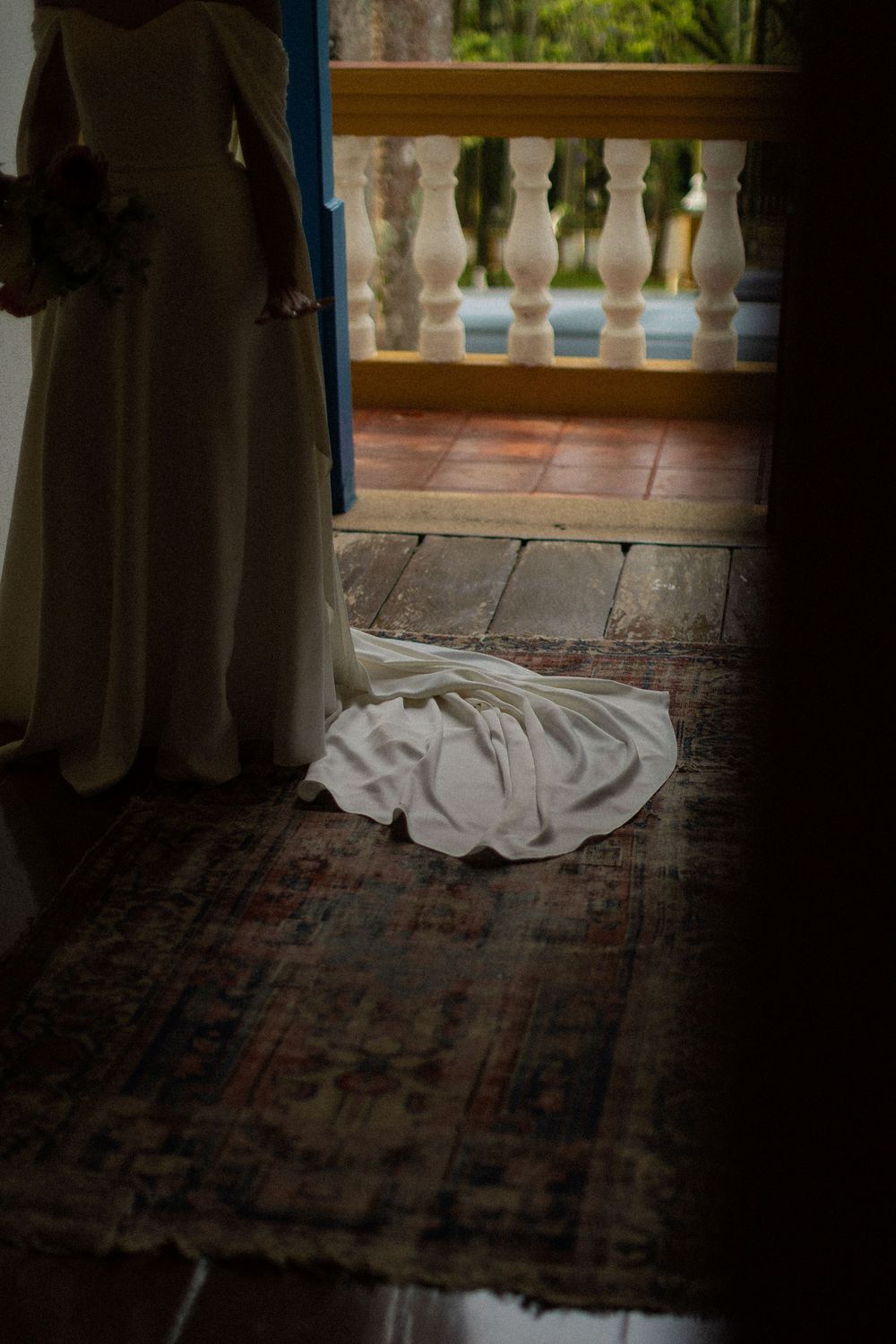 Bride in an elegant white gown stands in a doorway, her train spread across a vintage rug, with a balcony view beyond.