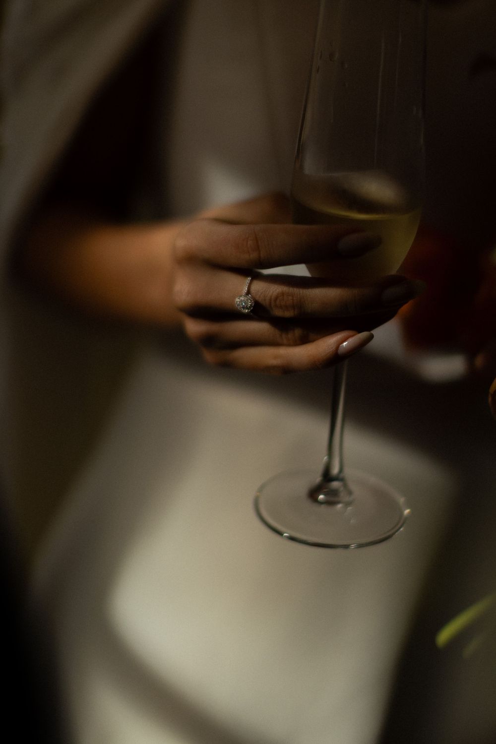 Woman's hand with a diamond ring and manicured nails holding a glass of white wine in soft, moody lighting.