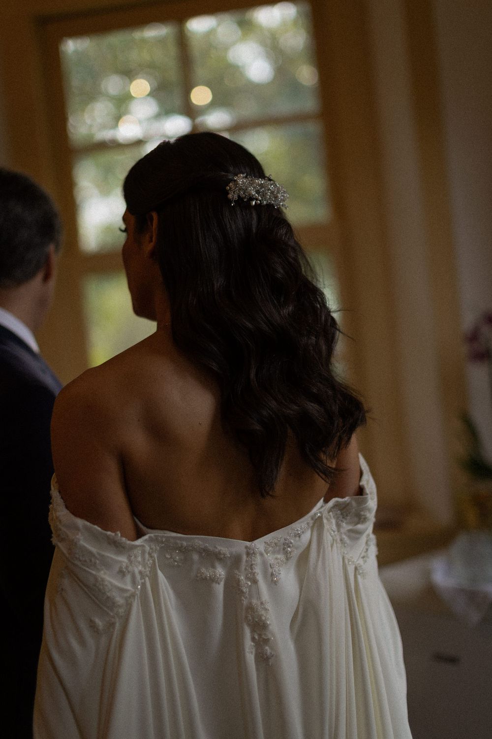 Bride seen from behind in an off-the-shoulder white lace gown with long wavy dark hair and a sparkling hairpiece beside a groom in a suit.