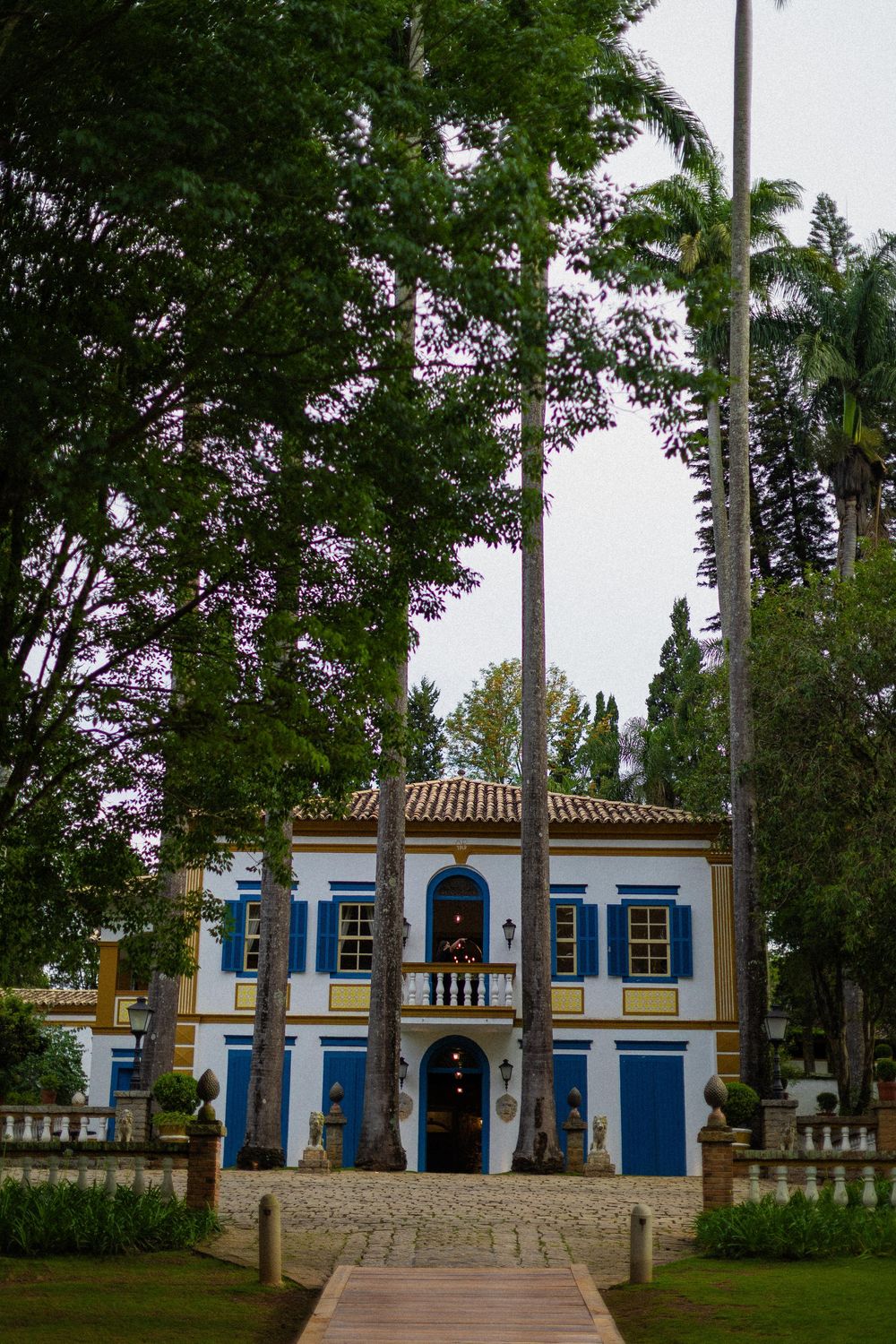 Colonial-style white manor with blue shutters and a tiled roof, framed by tall palm trees and lush gardens along a stone pathway.
