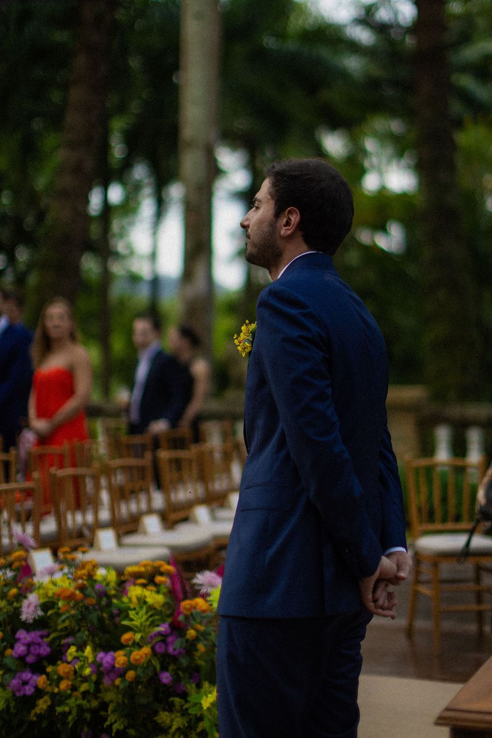 Groom in a navy suit with a yellow boutonniere stands at an outdoor garden wedding ceremony with guests and colorful flowers behind him.