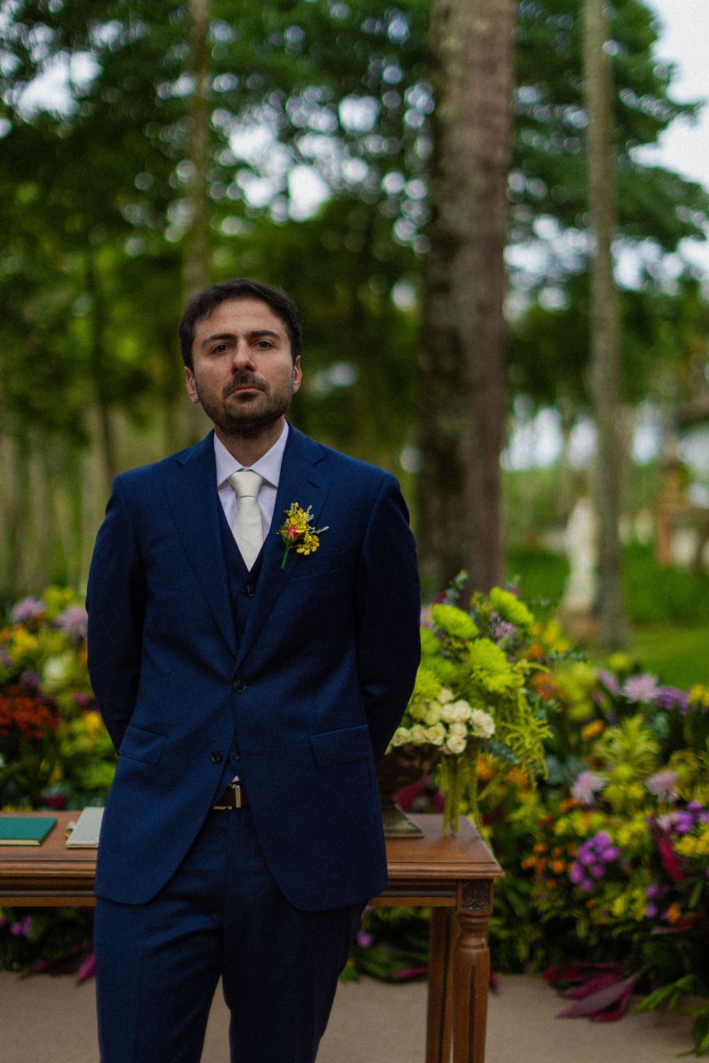 Bearded groom in a navy three-piece suit with a yellow boutonniere stands beside a wooden table and colorful flowers at an outdoor wedding.