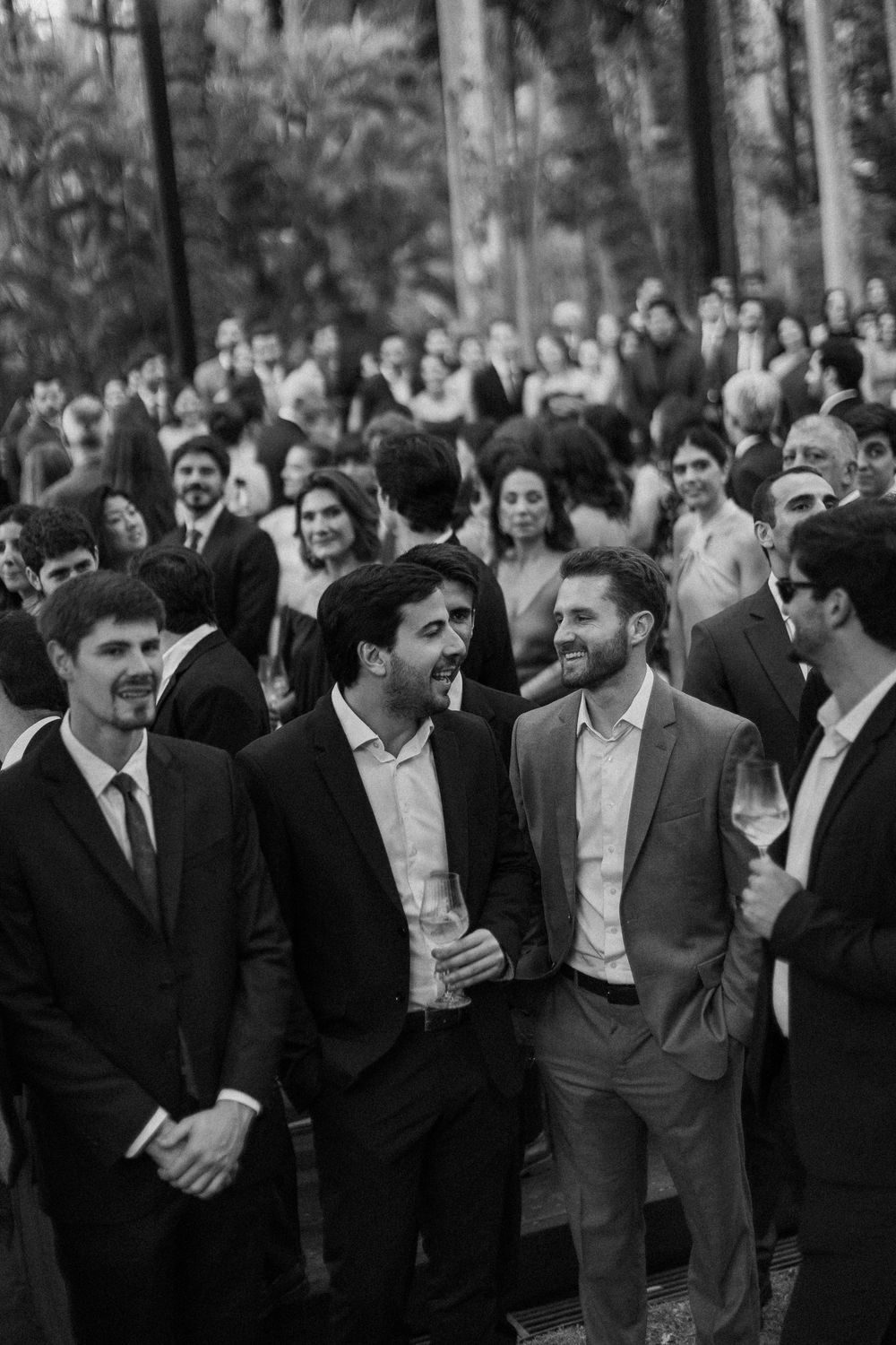 Black and white photo of smiling men in suits laughing together at an outdoor wedding reception with a large crowd of guests behind them.