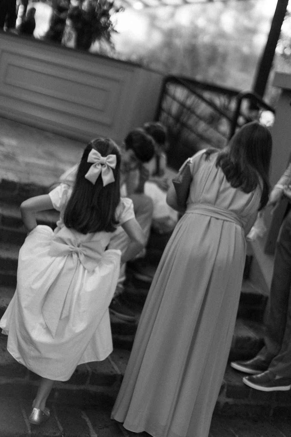 Black and white photo of a flower girl in a bow and tulle dress walking up brick steps beside a bridesmaid in a long gown.