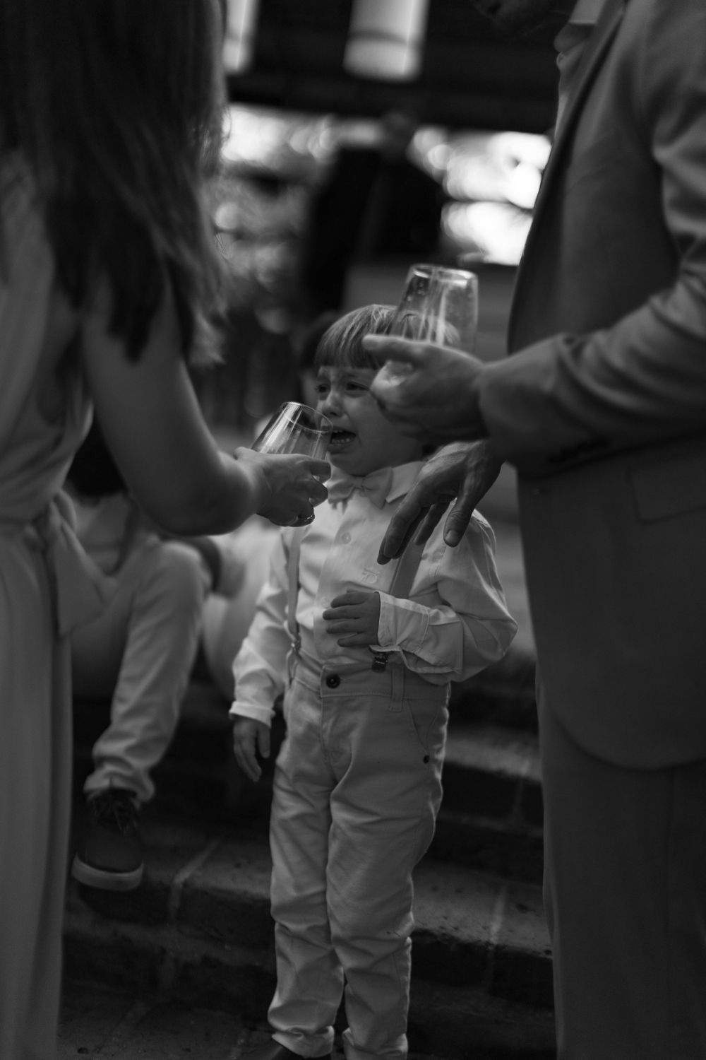 Black and white photo of a young boy in a bow tie and suspenders holding a glass while two adults hold glasses beside him on stone steps.