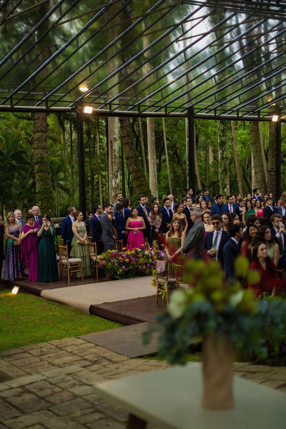 Wedding guests in formal attire stand during an outdoor ceremony beneath a glass pavilion surrounded by tall trees and lush forest.