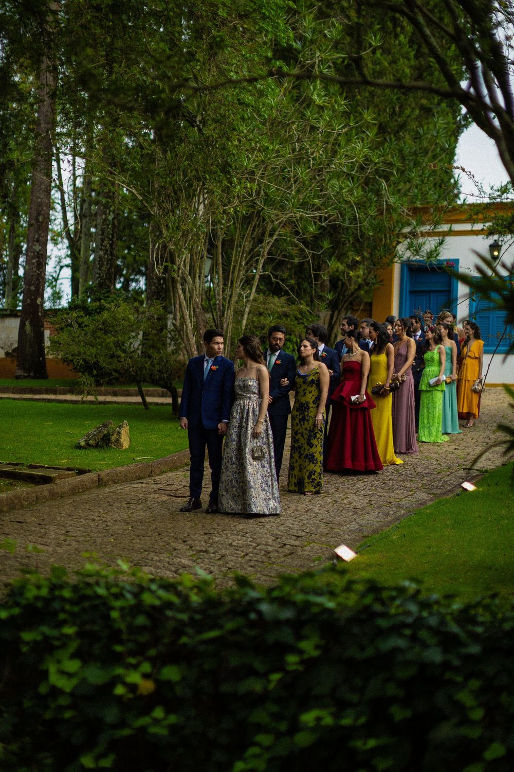 Wedding guests in colorful formal gowns and suits line up along a gravel garden path beneath tall trees beside a blue-windowed building.