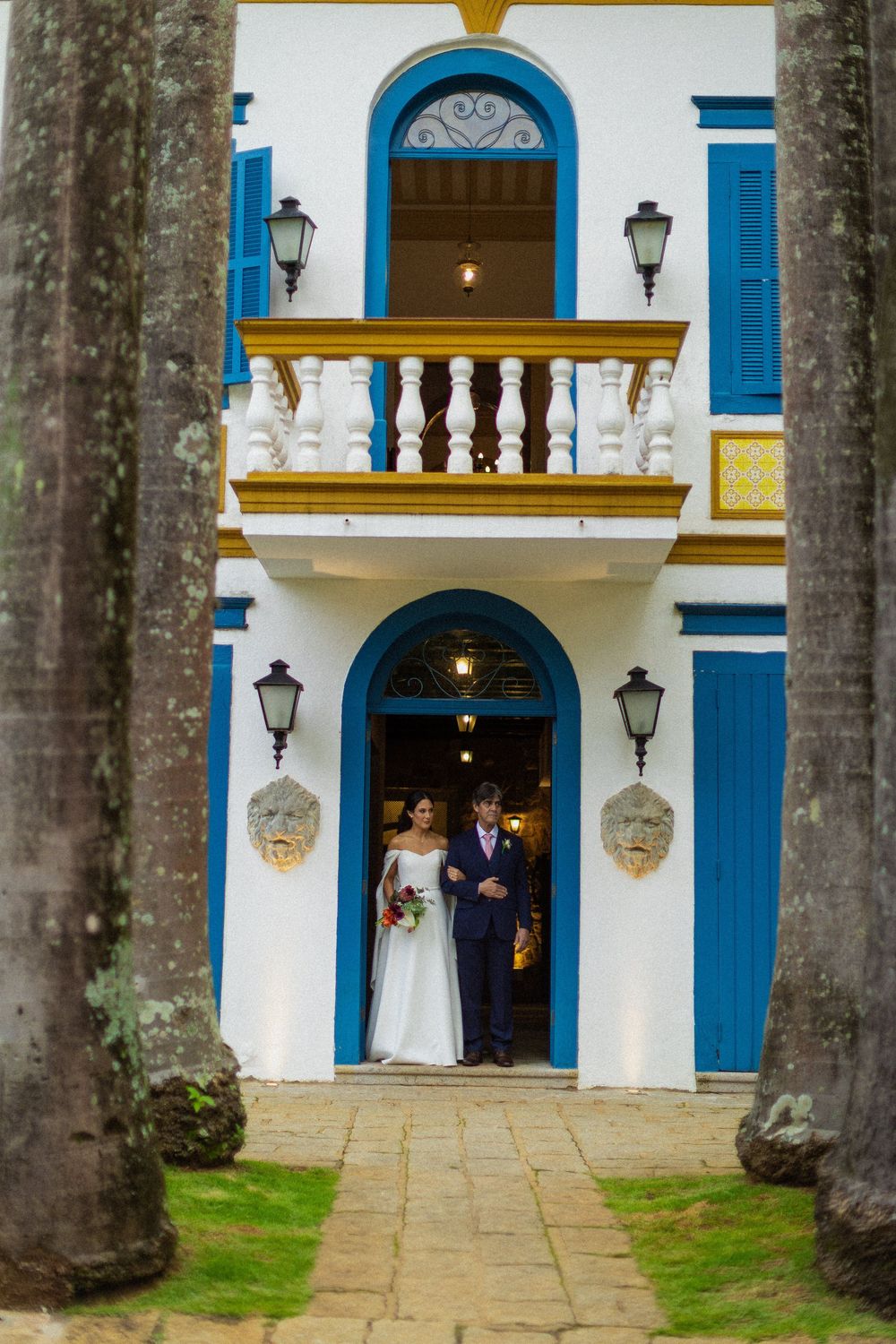 Bride in a white gown and groom in a navy suit stand in the doorway of a white colonial mansion with blue shutters, framed by tall trees.