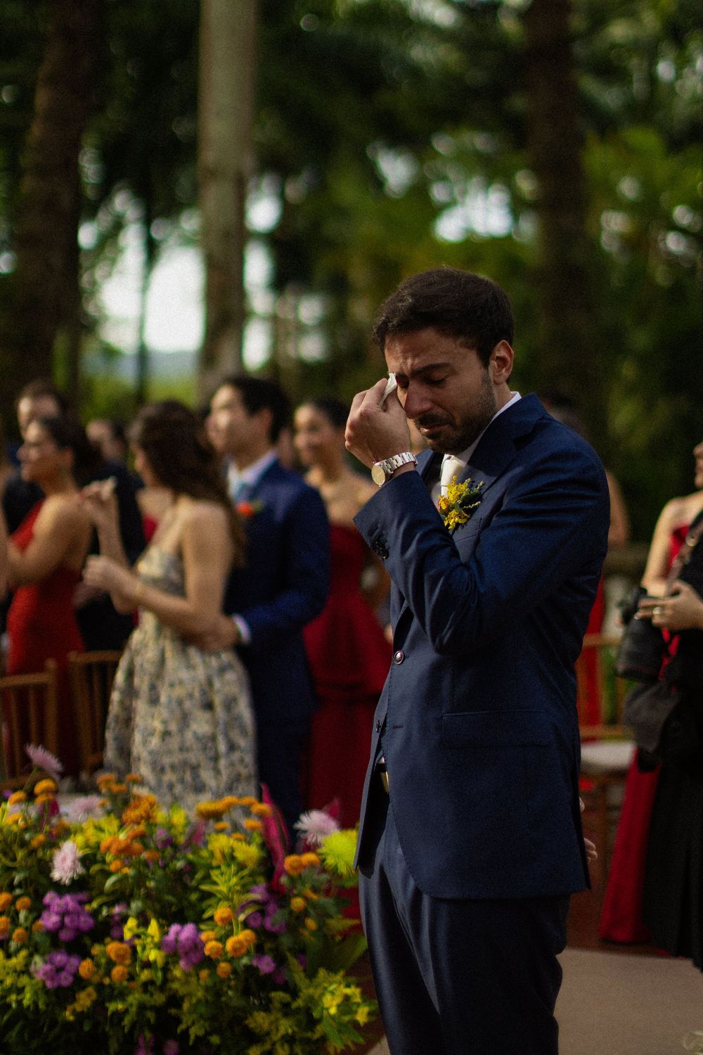 Emotional groom in a navy suit with a yellow boutonniere wipes away tears during an outdoor wedding ceremony as guests look on.