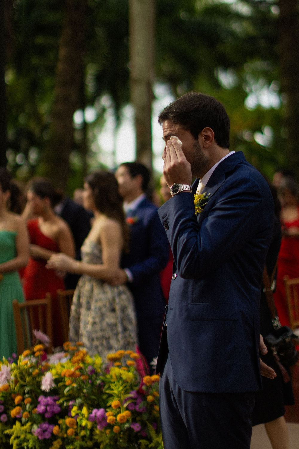 Emotional groom in a navy suit wipes his tears at an outdoor wedding ceremony as guests and colorful flowers fill the background.