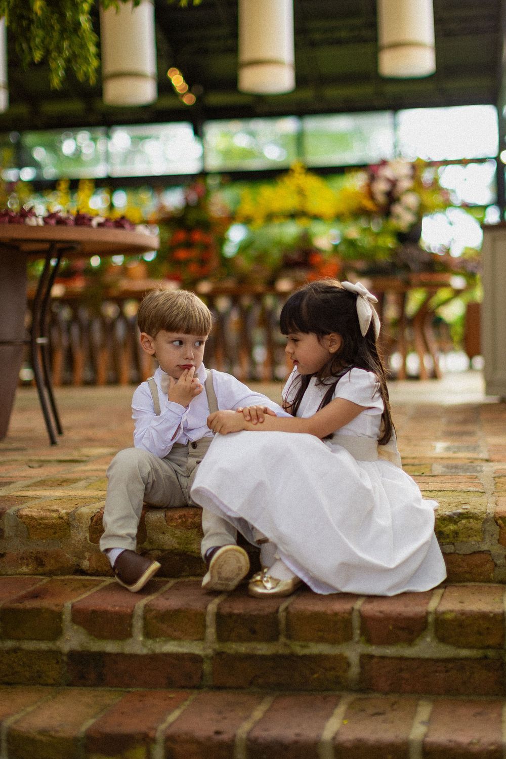 Young boy and girl in formal wedding attire sit close together on brick steps in a sunlit garden, the boy resting his chin on his hand.