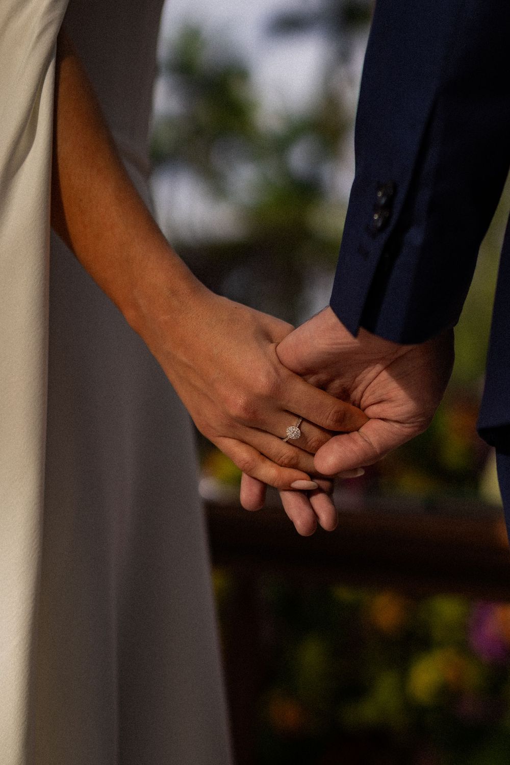 Close-up of a couple holding hands, the bride wearing a diamond engagement ring, with a blurred garden in the background.