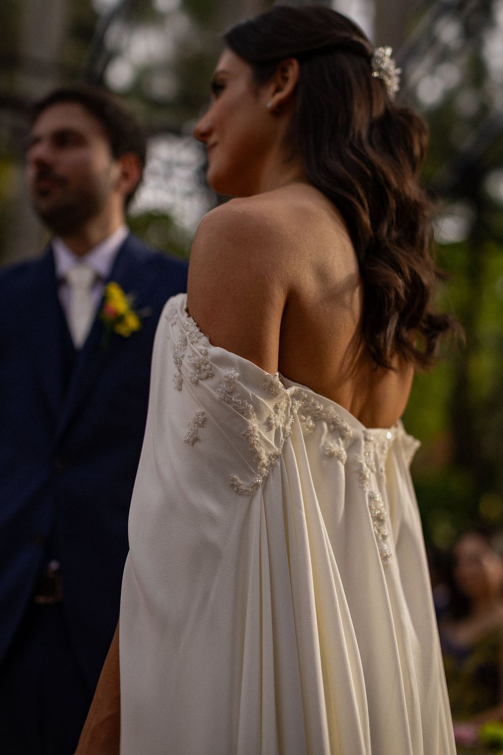 Bride in an off-the-shoulder white gown with beaded details and wavy hair stands beside a groom in a navy suit at an outdoor wedding.