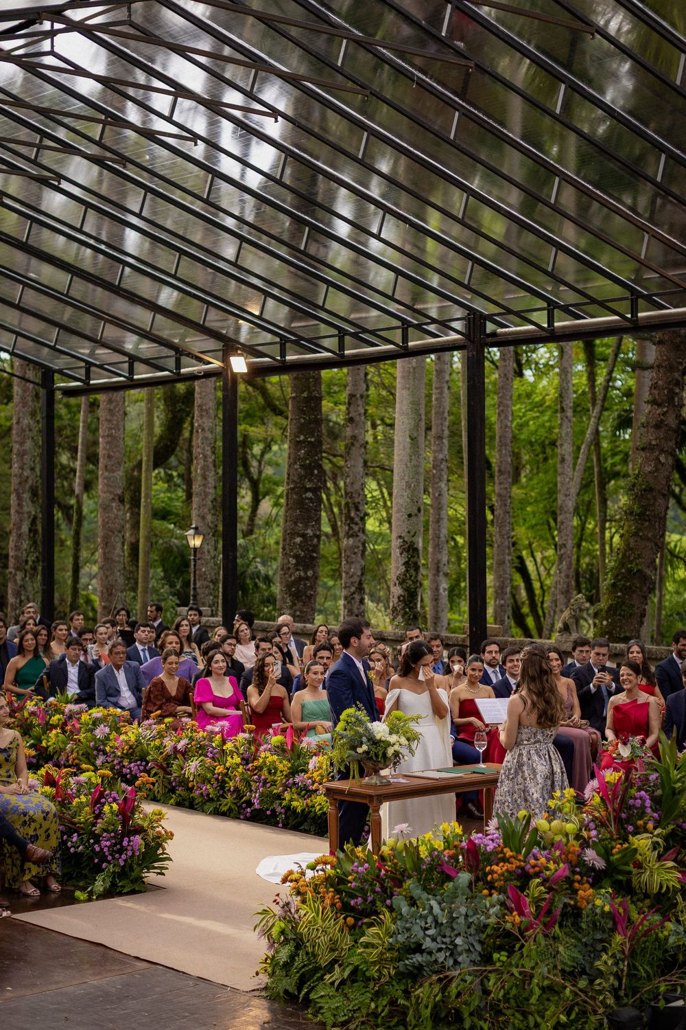 Wedding guests watch an outdoor ceremony under a glass-roofed pavilion surrounded by tall trees and colorful flower arrangements.