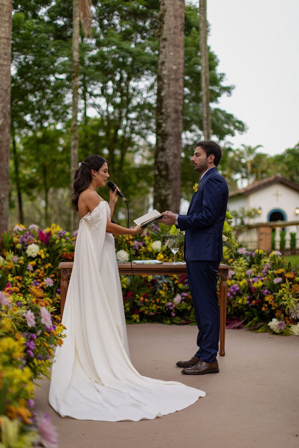 Bride in a flowing white gown speaks into a microphone as a groom in a navy suit holds a book at an outdoor garden wedding ceremony.