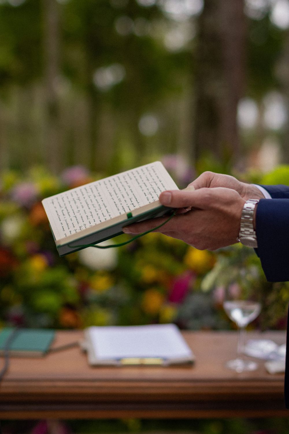Hands holding an open book during an outdoor wedding ceremony with colorful flowers, a wine glass and papers on a wooden table.