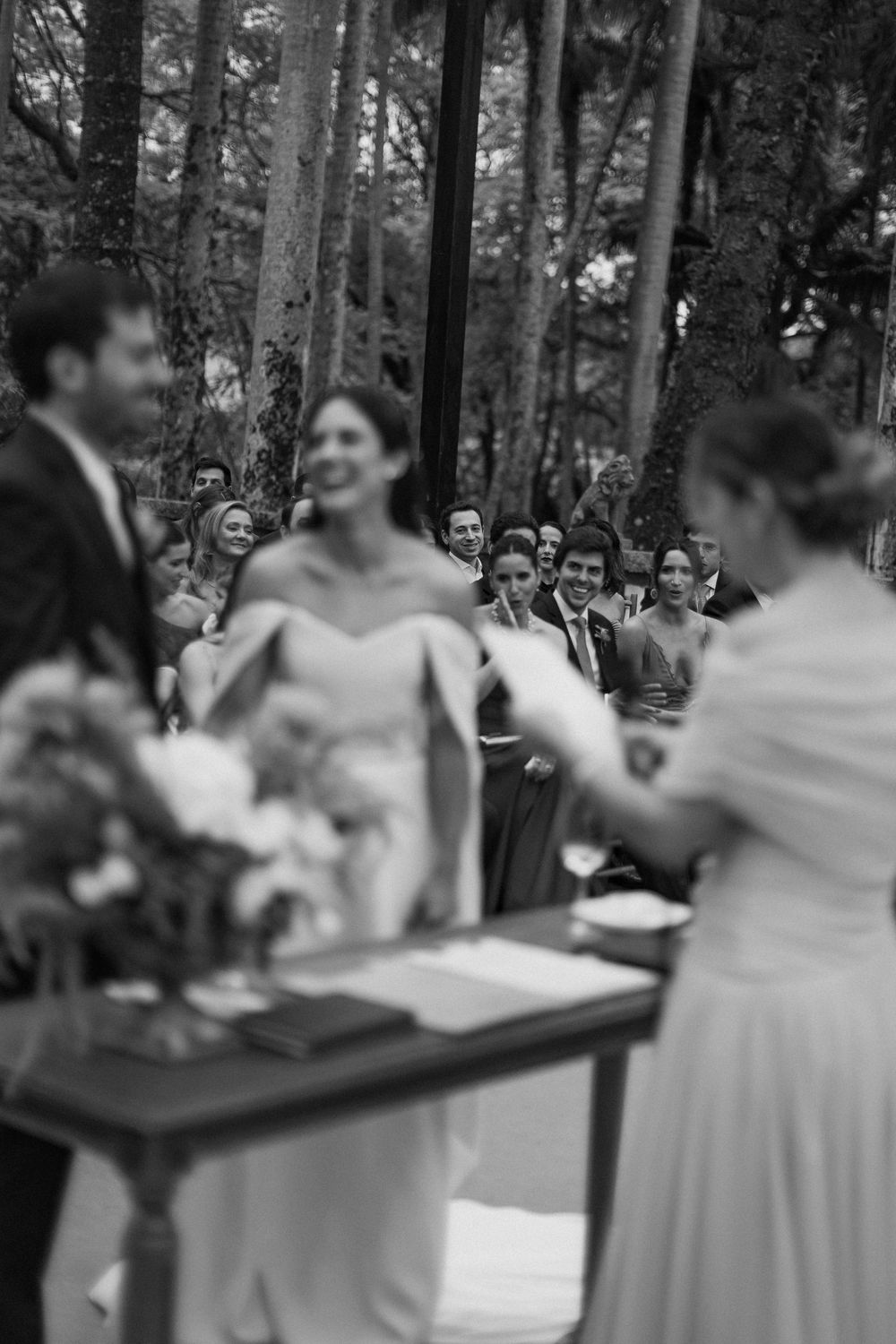 Black and white photo of a smiling bride in an off-shoulder gown at an outdoor forest wedding ceremony with guests watching behind her.