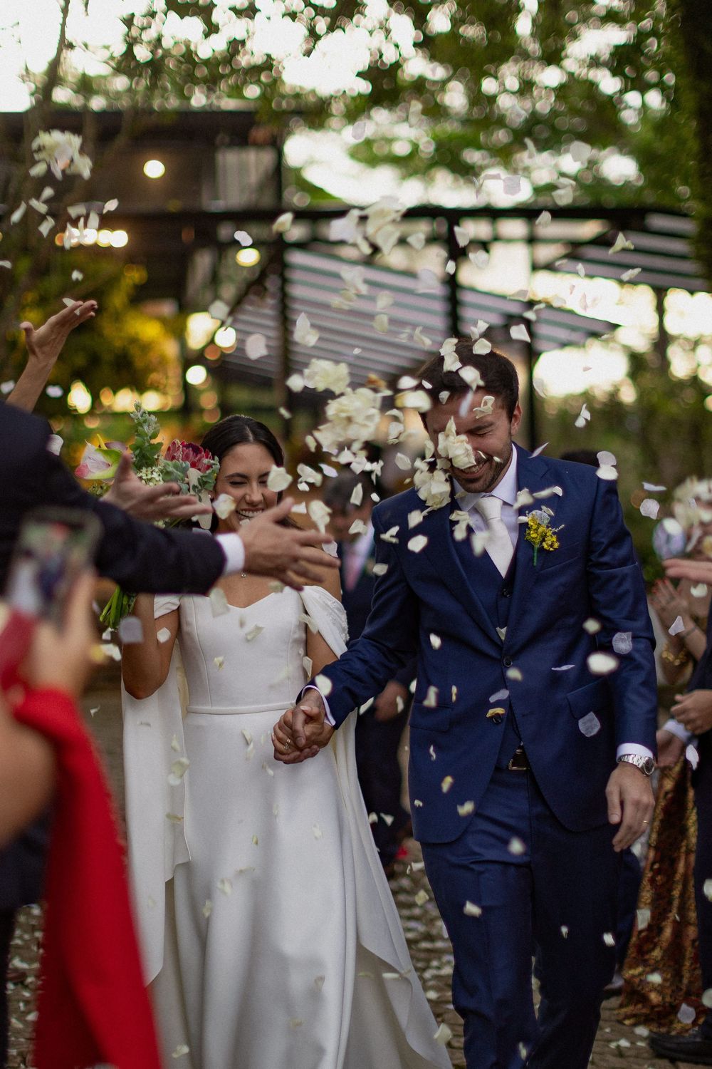 Smiling bride in a white gown and groom in a navy suit hold hands as guests toss petals during their outdoor wedding exit.