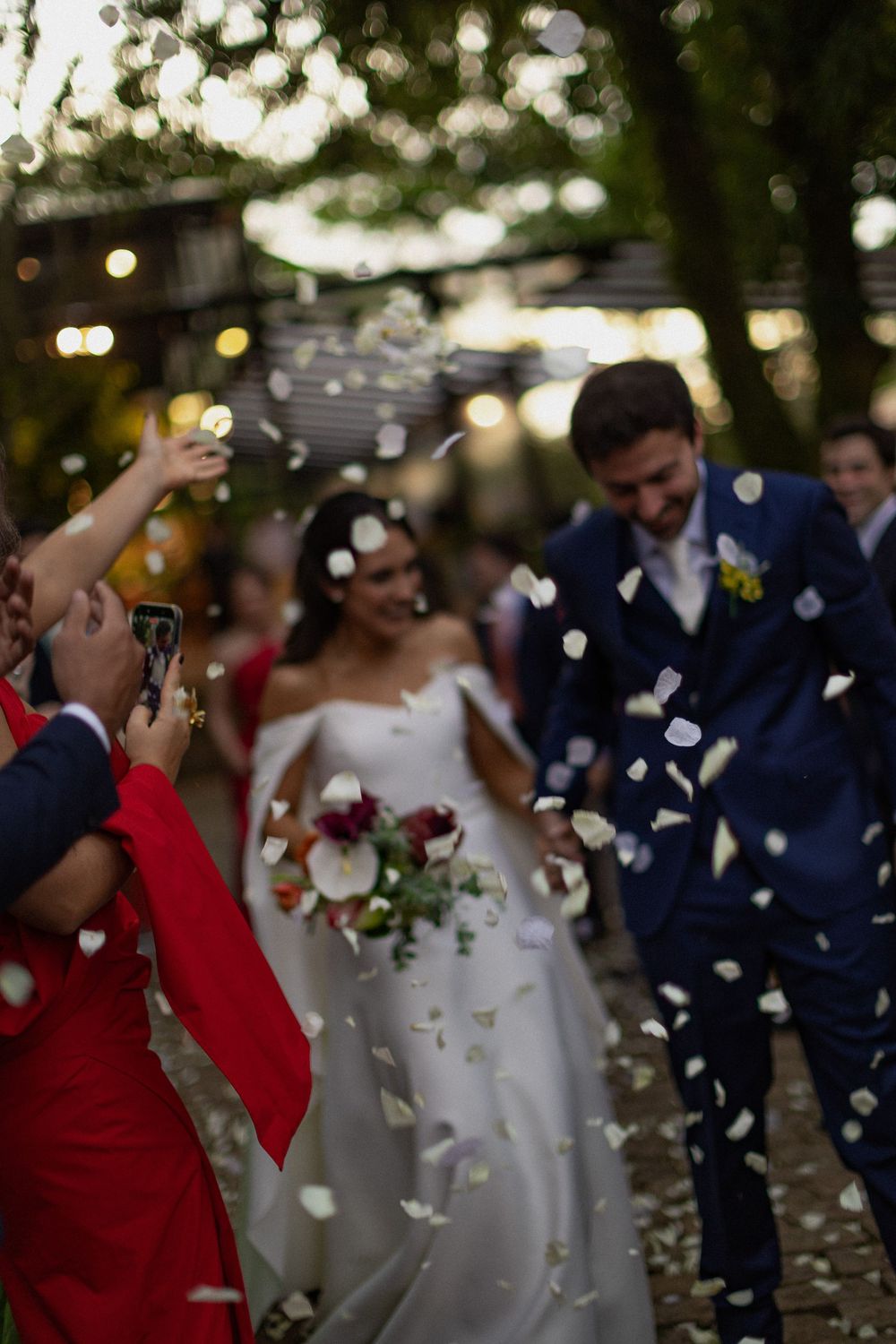 Bride in a white gown holding a bouquet and groom in a navy suit walk through falling petals as joyful wedding guests celebrate.