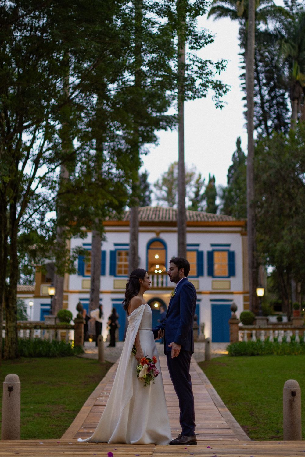 Bride in a flowing white gown holding a bouquet stands with her groom on a wooden walkway before a elegant colonial-style estate at dusk.