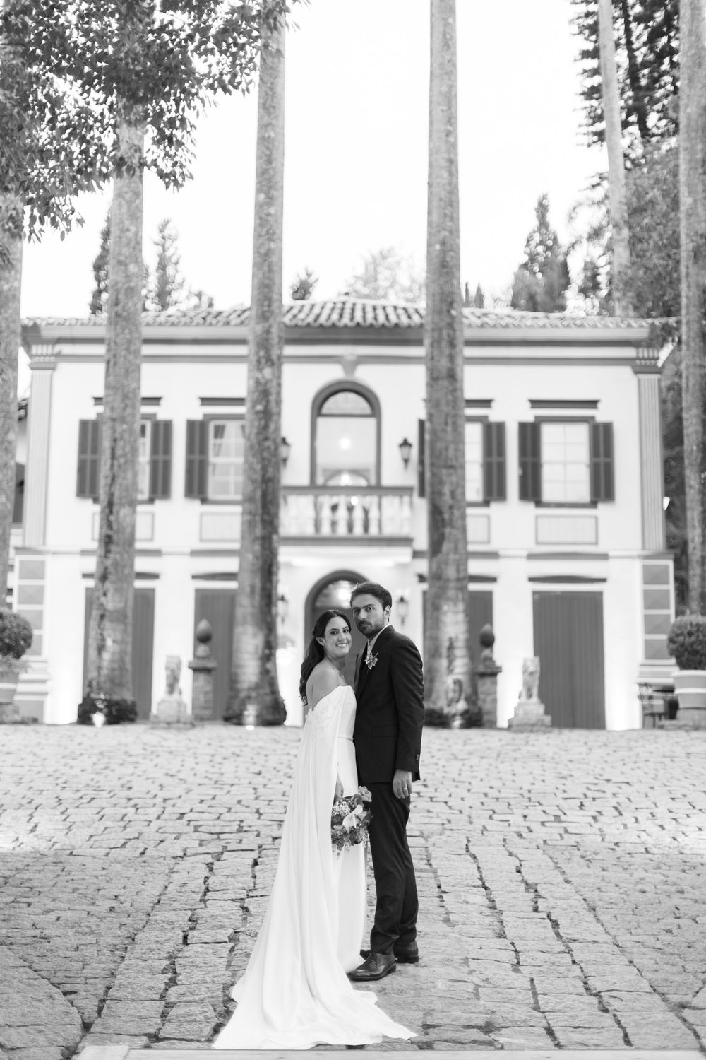 Black and white photo of a bride and groom embracing on a cobblestone courtyard in front of a historic manor lined with tall trees.
