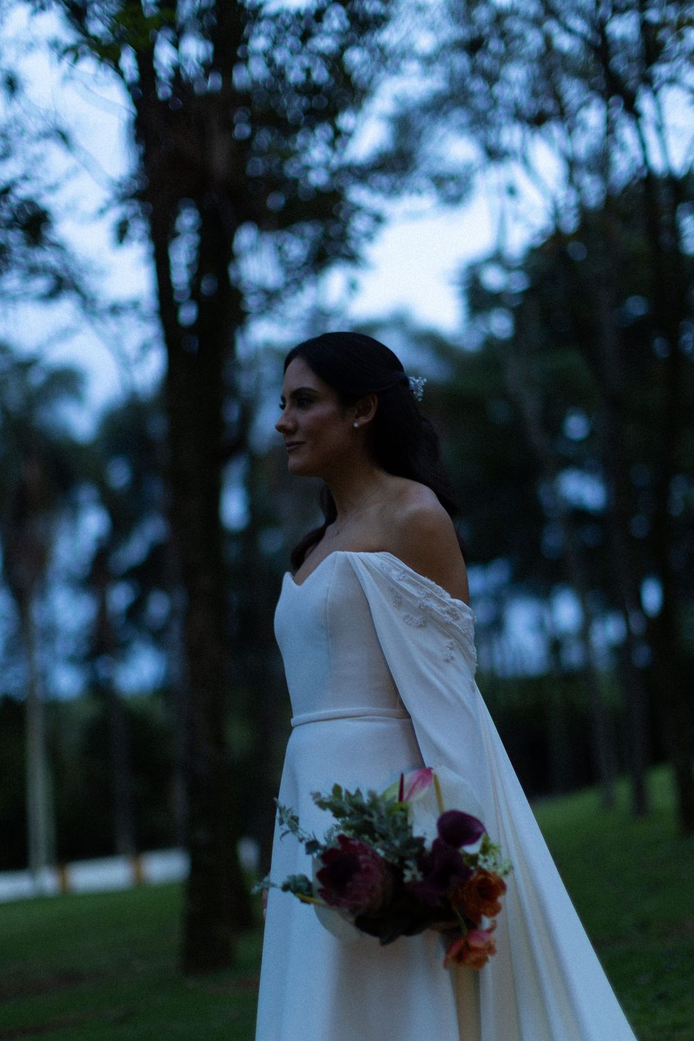 Bride in an off-the-shoulder white gown holds a lush burgundy and coral bouquet while standing in a tree-lined garden at dusk.