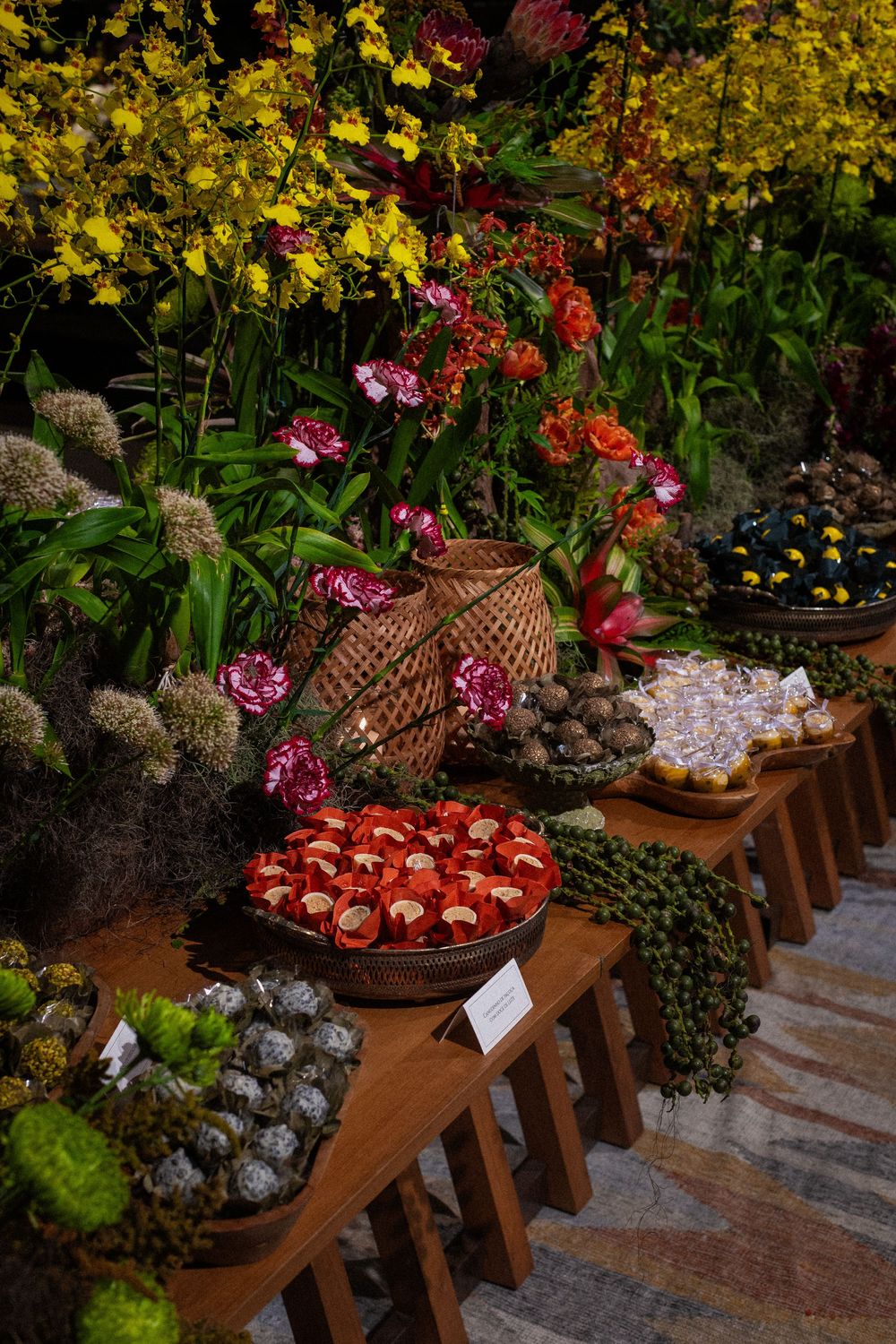 Colorful floral display with yellow orchids, pink and orange blooms, and baskets of red and gray ornamental plants on wooden tables.