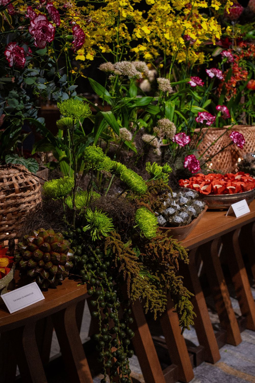 Floral display on wooden tables with pink carnations, yellow orchids, green foliage, woven baskets and a plate of sliced red fruit.