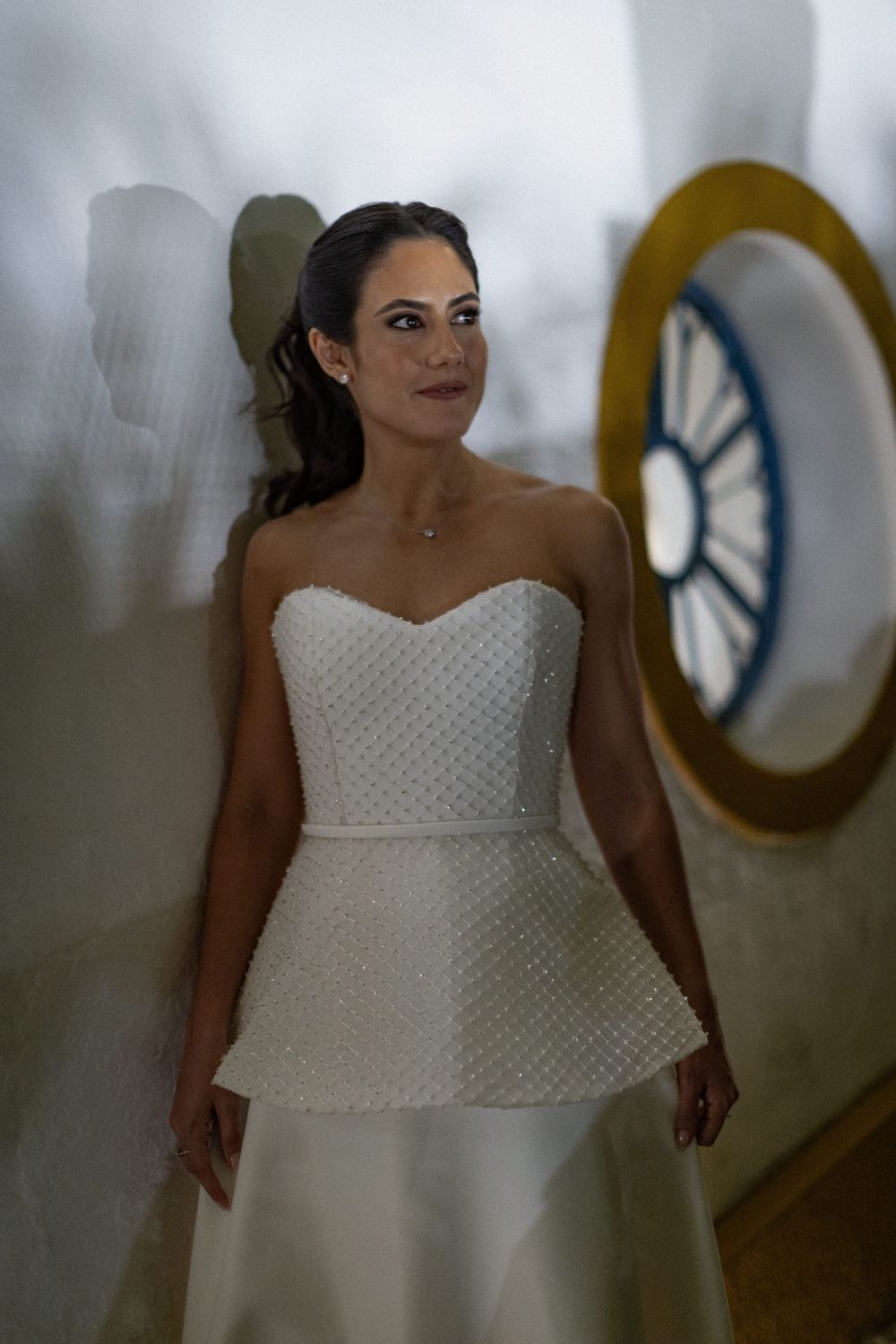 Bride in a strapless white beaded peplum gown with an updo stands beside a round porthole window, glancing to the side.
