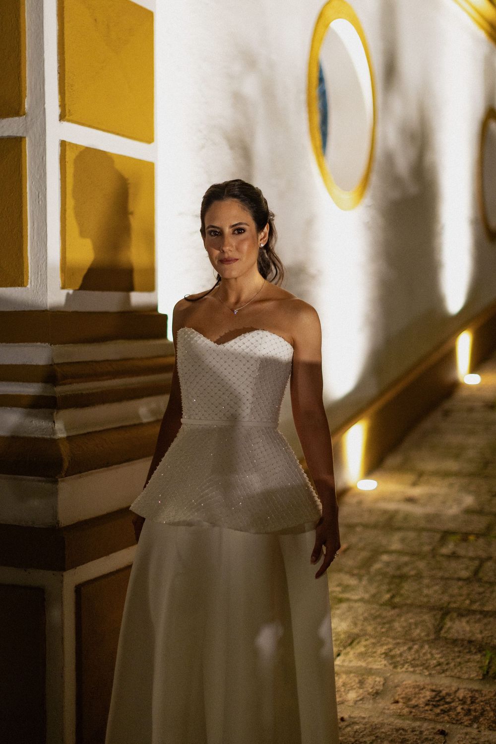 Bride in an elegant strapless white peplum wedding gown standing on a cobblestone path beside a yellow and white building at night.