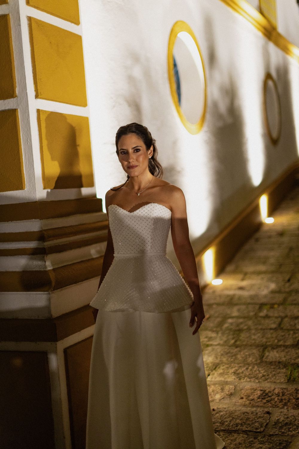 Bride in an elegant strapless white wedding gown with a peplum bodice standing on a cobblestone path beside a softly lit yellow-trimmed wall